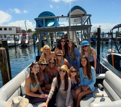 Group of smiling women in swimwear and sun hats aboard a pontoon boat at a sunny marina, blue sky and puffy clouds overhead.
