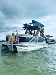 Cheerful group on a pontoon boat with stacked paddleboards, anchored in shallow green water near a floating tiki bar under an overcast sky