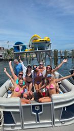 Group of friends in colorful bikinis cheering on a pontoon boat at a sunny marina with docks and inflatable water gear