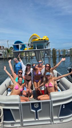 Group of friends in colorful bikinis cheering on a pontoon boat at a sunny marina with docks and inflatable water gear