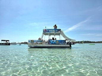 Sunlit pontoon party boat with a tall waterslide floating on crystal-clear turquoise shallow water under a bright blue sky, people lounging on deck and other boats in a calm coastal lagoon