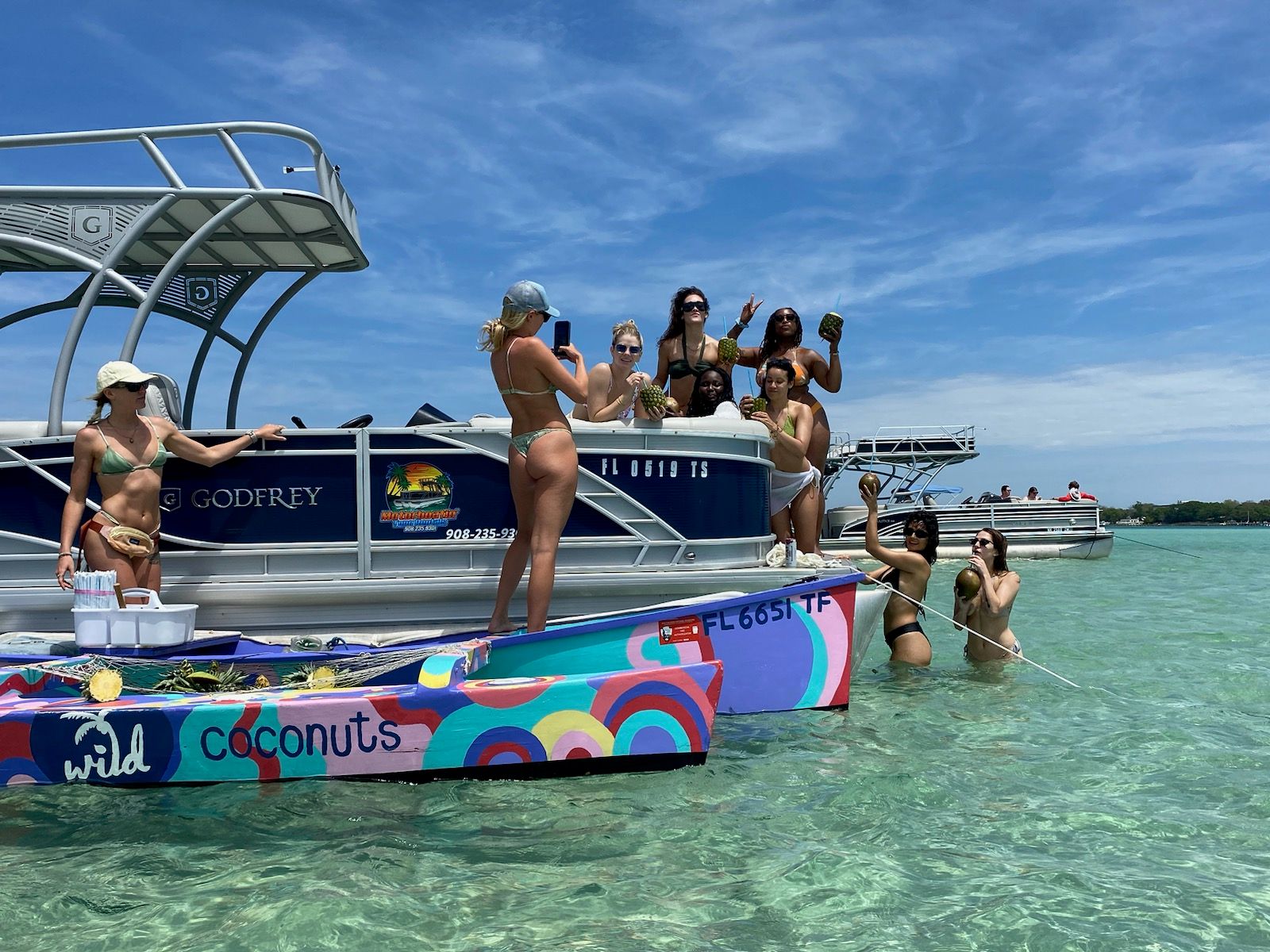 Group of friends on a pontoon and colorful small boat at a tropical sandbar, sunlit clear turquoise shallow water, women in bikinis holding coconuts under a bright blue sky.