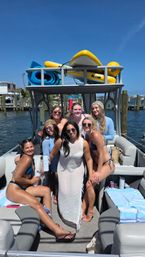 Group of friends in swimsuits smiling on a pontoon boat at a sunny marina, docked by wooden pilings with paddleboards and rolled beach mats on the upper rack — summer coastal boating scene.