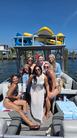 Group of friends in swimsuits smiling on a pontoon boat at a sunny marina, docked by wooden pilings with paddleboards and rolled beach mats on the upper rack — summer coastal boating scene.