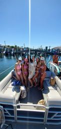 Smiling friends in colorful bikinis and a white cover-up posing on a pontoon boat at a sunny marina with clear blue sky and docked boats in the background, summer day on the water.
