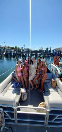 Smiling friends in colorful bikinis and a white cover-up posing on a pontoon boat at a sunny marina with clear blue sky and docked boats in the background, summer day on the water.
