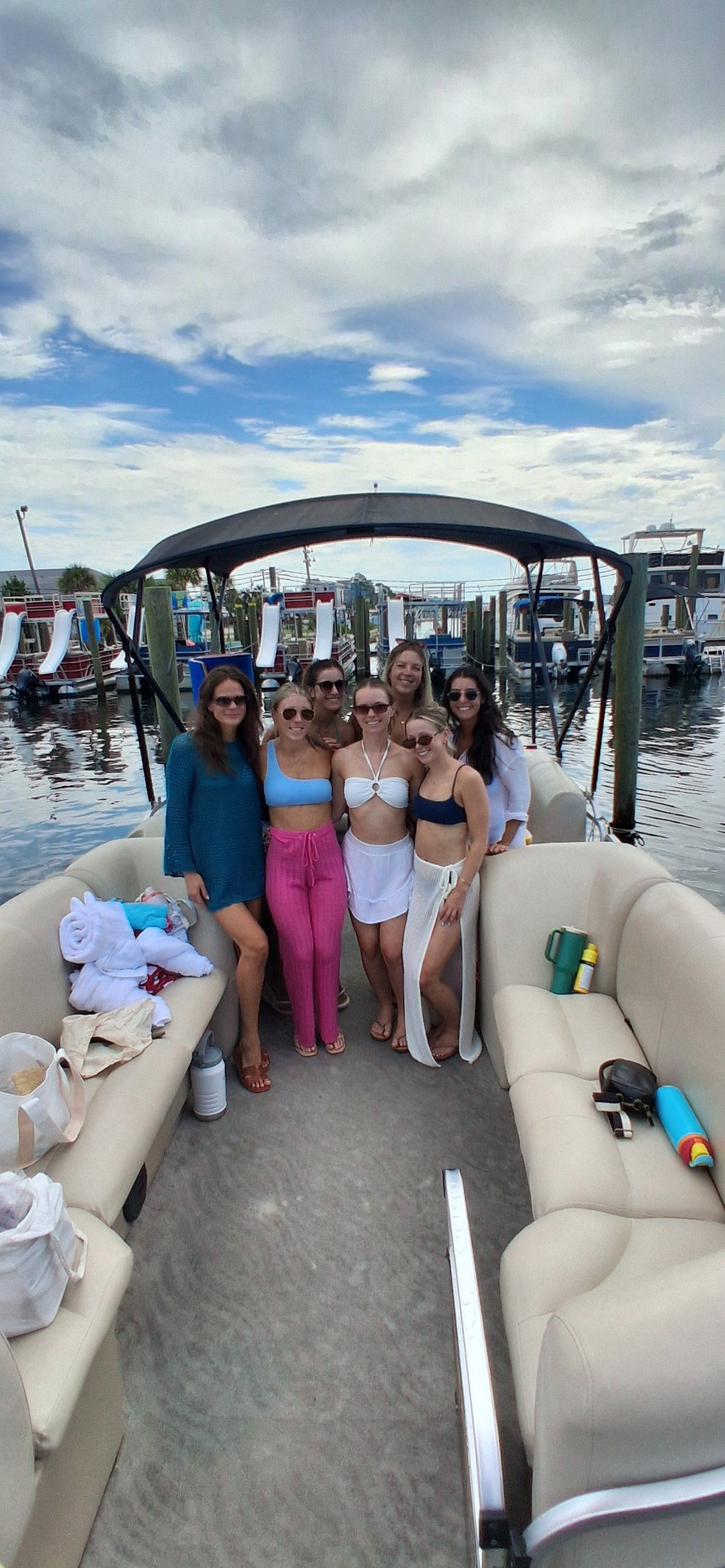 Seven friends in swimsuits smiling and posing on a pontoon boat at a marina dock, with other boats and a partly cloudy summer sky in the background