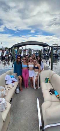 Seven friends in swimsuits smiling and posing on a pontoon boat at a marina dock, with other boats and a partly cloudy summer sky in the background