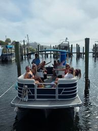 Group of friends smiling and waving on a pontoon boat docked at a coastal marina, palm trees and other boats in the sunny waterfront background.