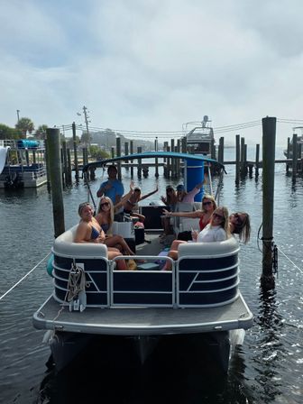 Group of friends smiling and waving on a pontoon boat docked at a coastal marina, palm trees and other boats in the sunny waterfront background.