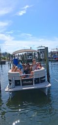 Group of people relaxing on a pontoon boat at a sunny marina dock with blue sky and calm water — a summertime boating scene.