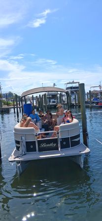 Group of people relaxing on a pontoon boat at a sunny marina dock with blue sky and calm water — a summertime boating scene.