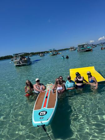 Group of people enjoying a sunny sandbar: six women standing in crystal-clear turquoise water around a paddleboard and bright yellow floating mat, with pontoon boats and swimmers anchored nearby under a vivid blue sky.