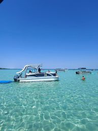Pontoon boat with a white slide anchored in crystal-clear turquoise shallow water near a sandbar, people wading and other boats under a bright blue sky