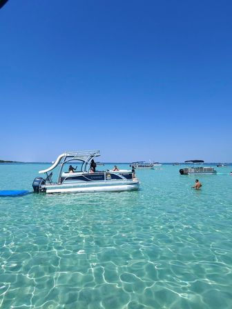 Pontoon boat with a white slide anchored in crystal-clear turquoise shallow water near a sandbar, people wading and other boats under a bright blue sky