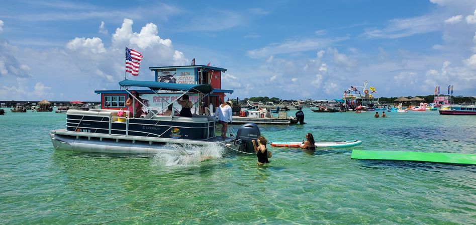 Festive pontoon boat flying an American flag in shallow turquoise coastal water with people swimming, paddleboarding, and colorful party boats under a sunny blue sky.