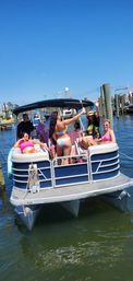 Friends partying on a blue pontoon boat at a sunny marina, colorful bikinis and raised drinks with dock pilings and clear blue sky