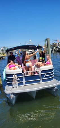 Friends partying on a blue pontoon boat at a sunny marina, colorful bikinis and raised drinks with dock pilings and clear blue sky