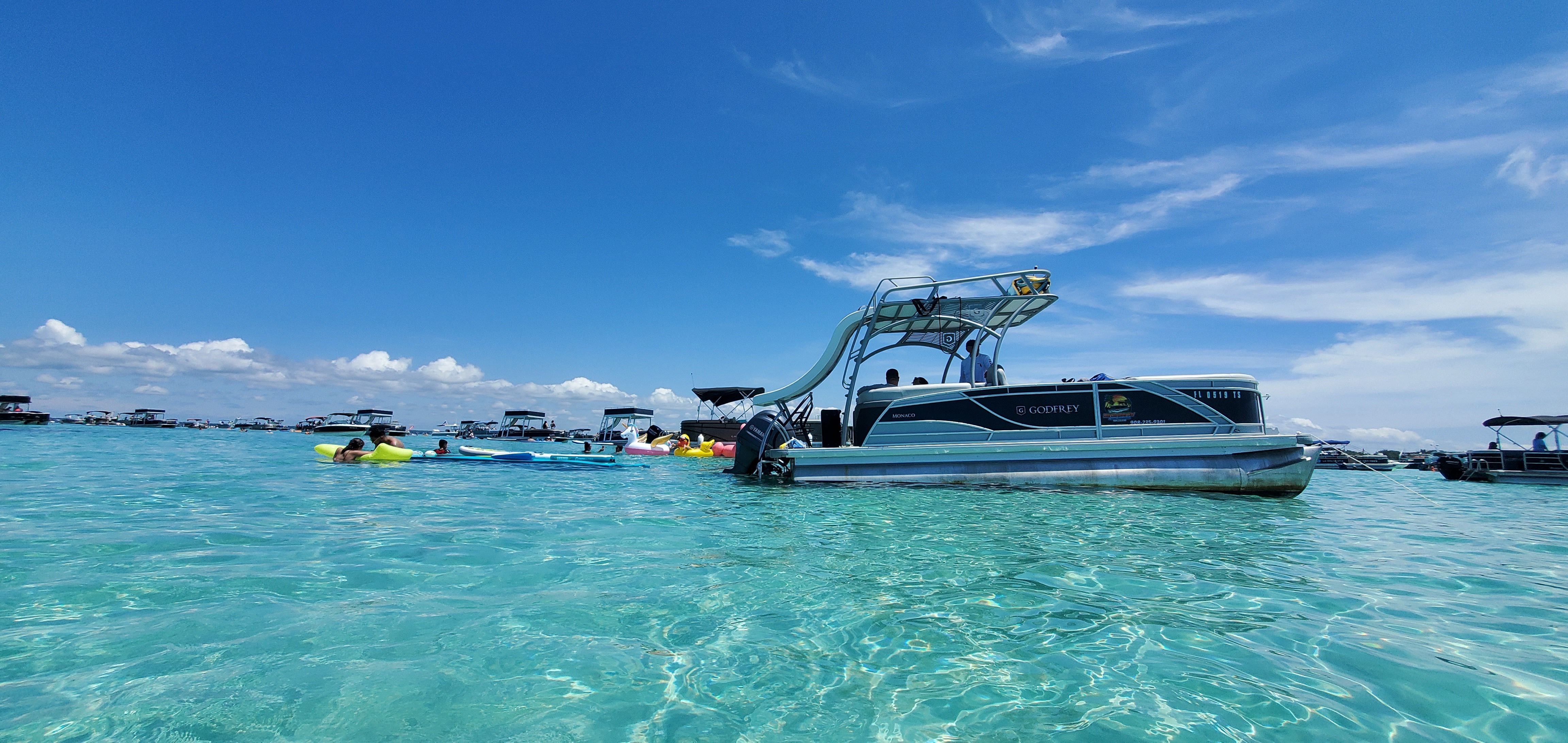 Pontoon boat with waterslide anchored in clear turquoise shallow water, people on colorful inflatables and nearby boats under a bright blue sky