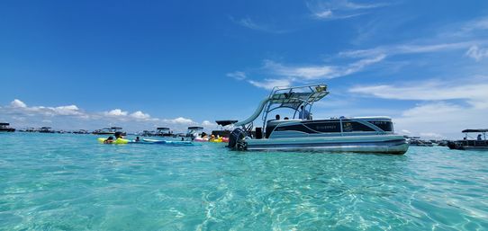 Pontoon boat with waterslide anchored in clear turquoise shallow water, people on colorful inflatables and nearby boats under a bright blue sky