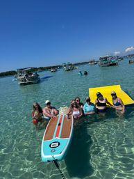 Group of people standing in crystal-clear shallow turquoise water around a striped paddleboard and bright yellow floating mat, with anchored pontoon boats and swimmers nearby under a sunny blue sky at a popular sandbar