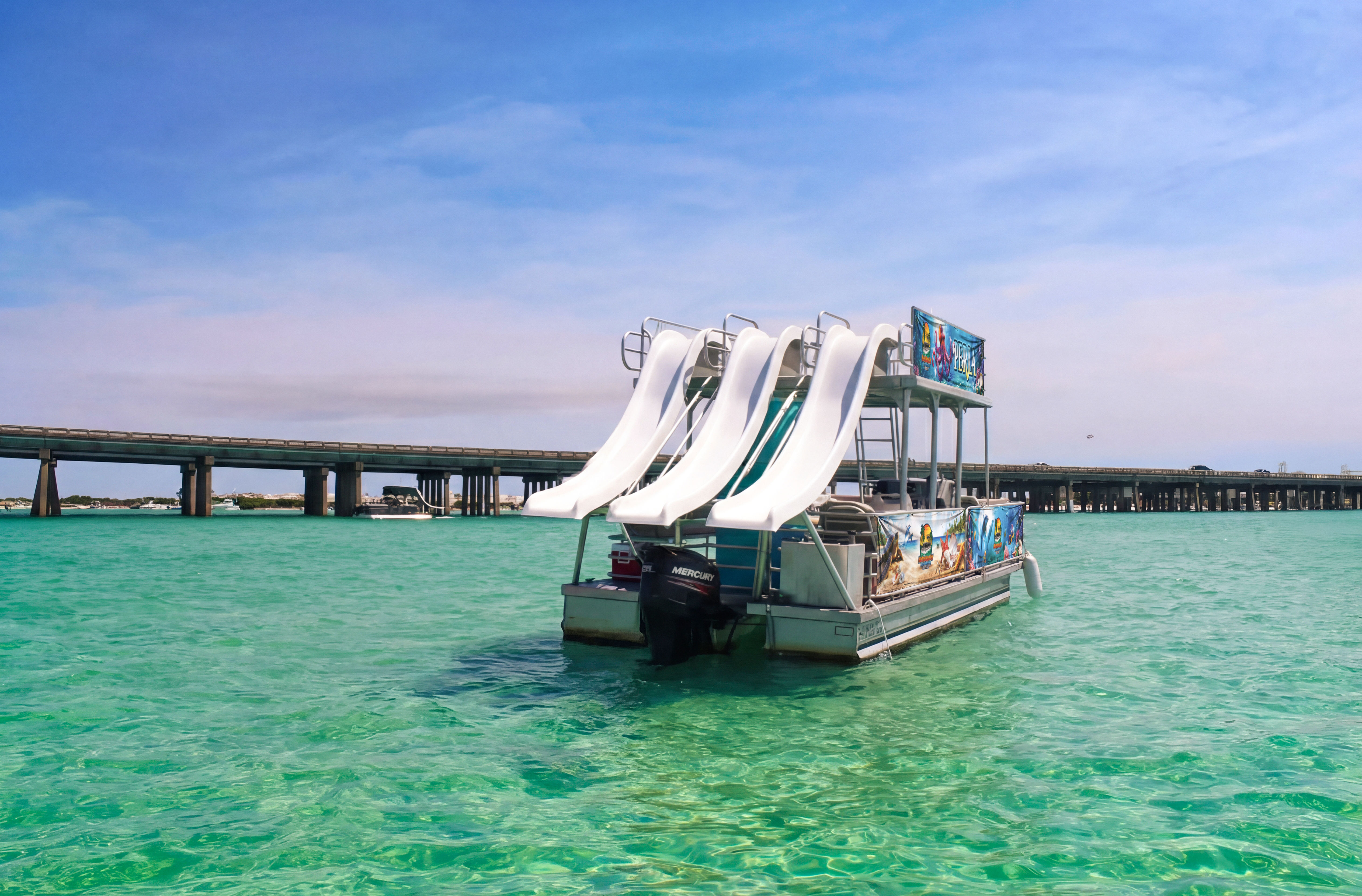 Pontoon party boat with three white water slides floating in clear turquoise coastal waters near a low concrete bridge under a sunny blue sky.