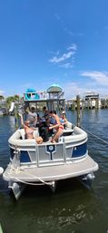 Smiling group on a blue-and-gray pontoon boat docked at a sunny marina, people in swimsuits waving under a clear blue sky
