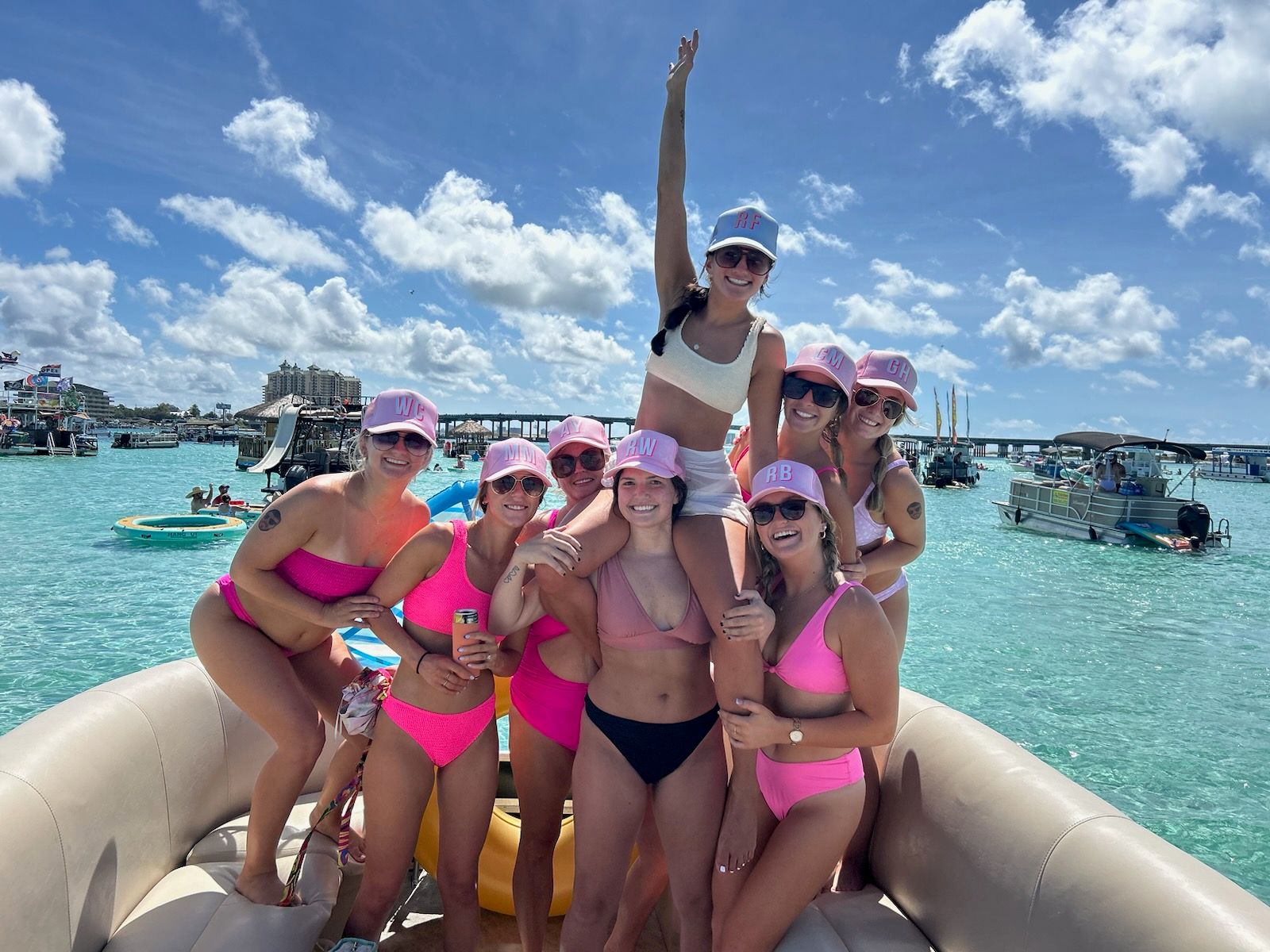 Group of friends in pink bikinis and matching caps smiling and posing on an inflatable boat in turquoise water with other boats and a sunny blue sky — lively boat party vibe.