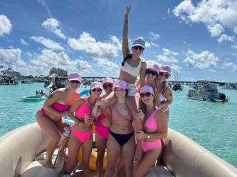 Group of friends in pink bikinis and matching caps smiling and posing on an inflatable boat in turquoise water with other boats and a sunny blue sky — lively boat party vibe.
