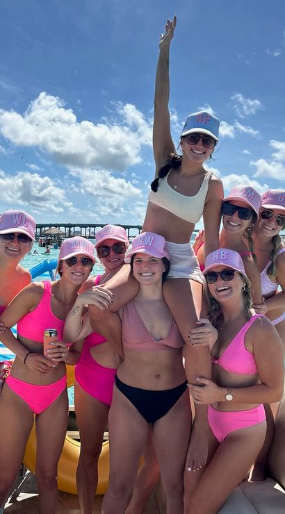 Group of friends in pink bikinis and matching caps smiling and posing on an inflatable boat in turquoise water with other boats and a sunny blue sky — lively boat party vibe.