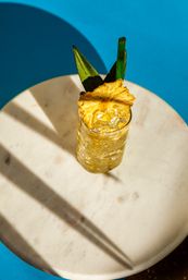Sunlit tropical pineapple cocktail in a tall glass with crushed ice, dried pineapple slice, green straw and leaves on a round marble table against a bright blue background.