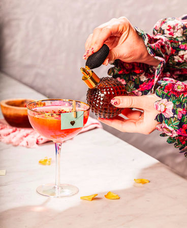 Vintage perfume atomizer spritzing a pink rose-petal cocktail in a coupe glass with a tiny heart note clipped to the rim and scattered petals on a marble tabletop.