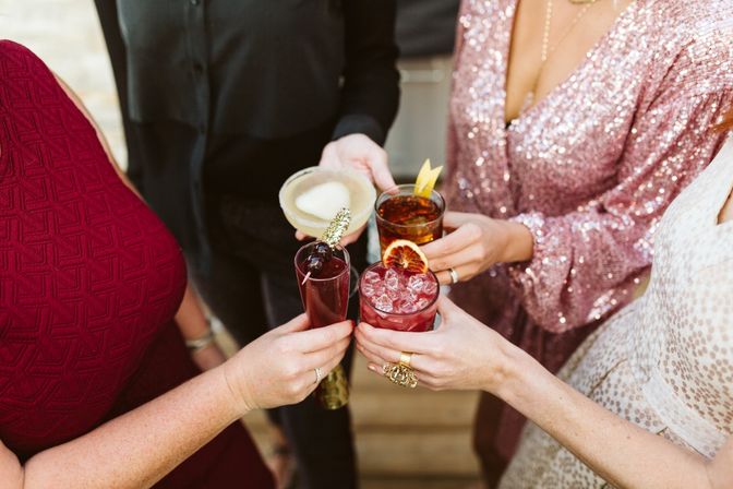 Four friends toasting colorful cocktails at a festive party — hands holding a salt-rim margarita, amber cocktail, iced red drink with dried orange garnish, and a cherry-topped flute; sparkly and burgundy party dresses visible.