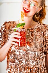 Smiling person in rose-gold sequin top sipping a red iced cocktail topped with mint and a honey dipper garnish.