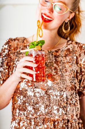 Smiling person in rose-gold sequin top sipping a red iced cocktail topped with mint and a honey dipper garnish.