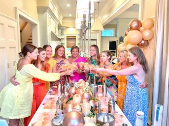 Group of women in bright summer dresses toasting cocktails over a decorated kitchen island with rose-gold balloons, metallic shakers, and festive table decor.