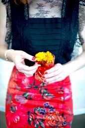 Close-up of a person in a black lace top and vibrant red floral skirt holding a red glass with bright yellow marigold flowers, red manicured nails and a gold bracelet visible.