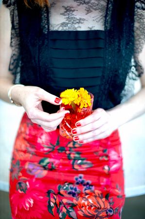 Close-up of a person in a black lace top and vibrant red floral skirt holding a red glass with bright yellow marigold flowers, red manicured nails and a gold bracelet visible.