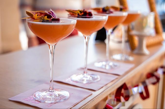 Row of peach-pink craft cocktails in coupe glasses topped with edible flowers and citrus slices, served on pink napkins along a wooden bar — elegant brunch or cocktail-hour presentation.