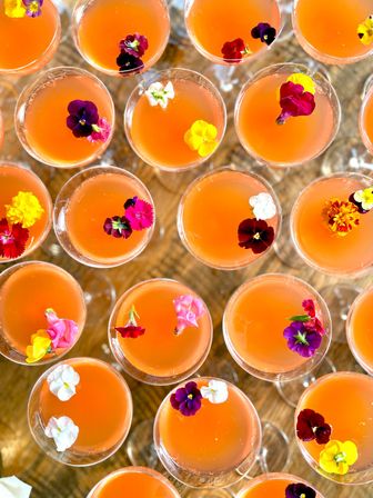 Overhead view of vibrant orange cocktails in coupe glasses, each topped with colorful edible flowers on a wooden table — bright summer party drink styling.