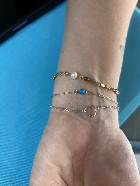 Close-up of a wrist wearing three delicate bracelets: a gold scalloped chain with a small pearl, a thin gold chain with a blue stone, and a silver chain with an open heart charm, set against bright turquoise fabric.