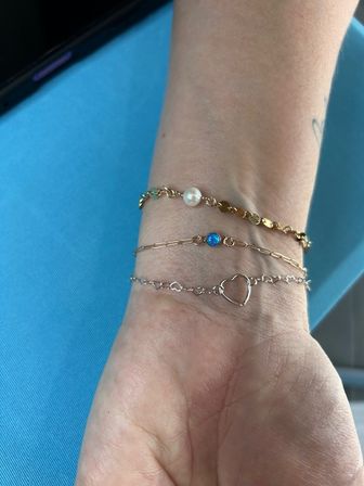Close-up of a wrist wearing three delicate bracelets: a gold scalloped chain with a small pearl, a thin gold chain with a blue stone, and a silver chain with an open heart charm, set against bright turquoise fabric.