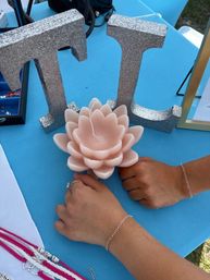 Two hands wearing delicate silver chain bracelets rest on a bright turquoise vendor table next to a blush pink lotus-shaped candle and sparkly silver decorative letters — jewelry display at an outdoor craft market.
