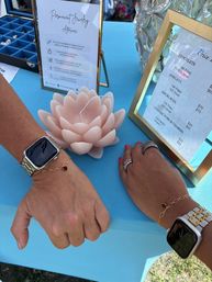 Two wrists wearing square smartwatches, gold chain bracelets and rings displayed on a bright turquoise vendor table with a pale pink lotus candle, framed price signs and a jewelry tray at an outdoor craft market