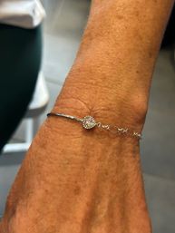Close-up of a tanned wrist wearing a delicate silver-tone bracelet with a heart-shaped pavé crystal charm against a soft indoor background