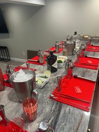Marble kitchen island set up for a cocktail-making session with red bar mats, stainless steel shakers and jiggers, ice bucket, glassware and bowls of cucumber slices and fresh herbs — modern home mixology station.