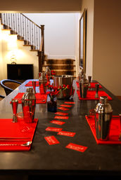 Modern home bar setup on a long kitchen island featuring stainless steel cocktail shakers, jiggers and ice bucket, bright red placemats and flower accents, small red cards scattered down the tabletop, warm-lit interior with a staircase in the background