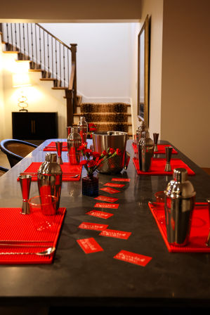Modern home bar setup on a long kitchen island featuring stainless steel cocktail shakers, jiggers and ice bucket, bright red placemats and flower accents, small red cards scattered down the tabletop, warm-lit interior with a staircase in the background