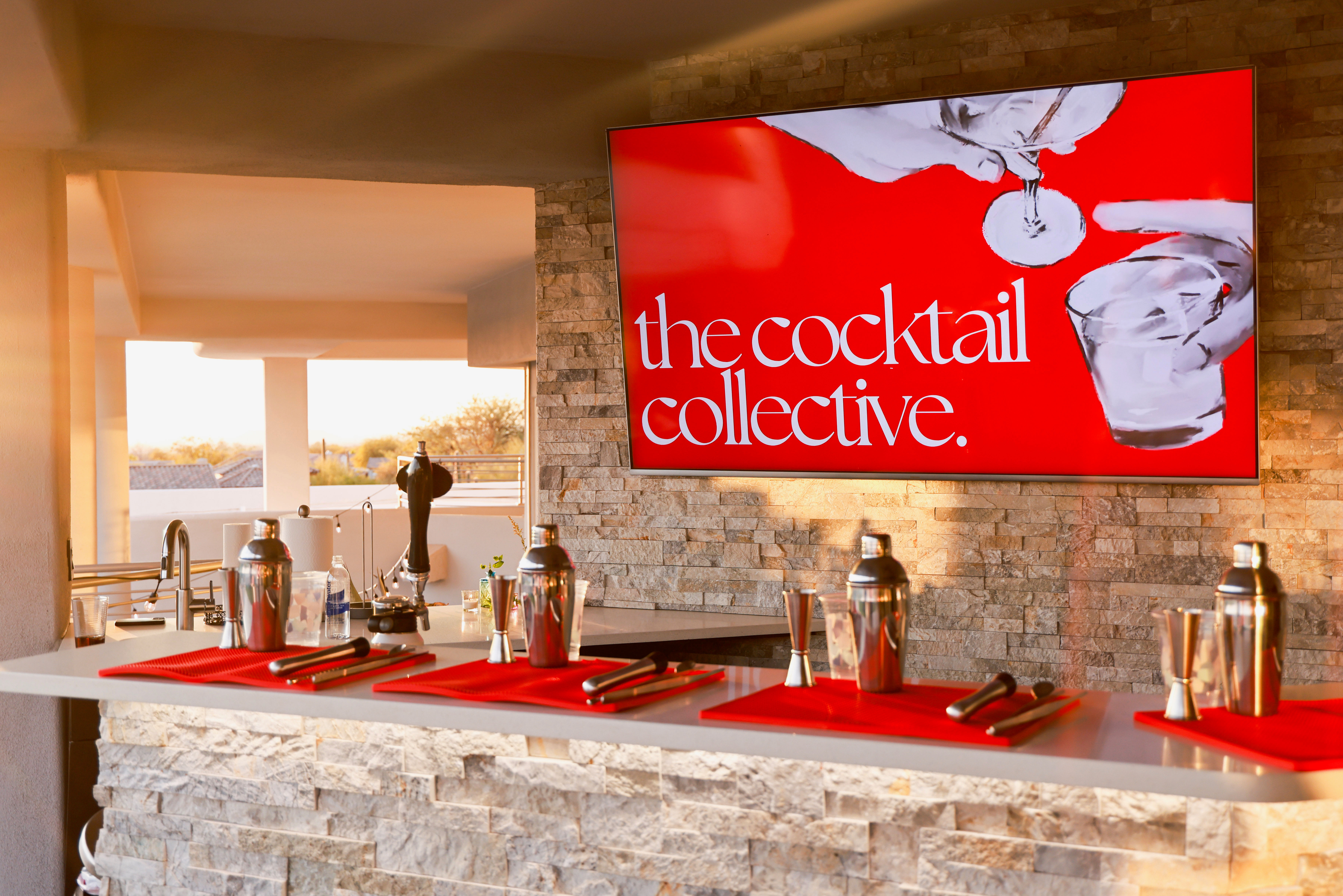 Sunset-lit outdoor patio bar with stone counter and a large red cocktail sign, lined with stainless-steel shakers, jiggers and bar tools on red mats.