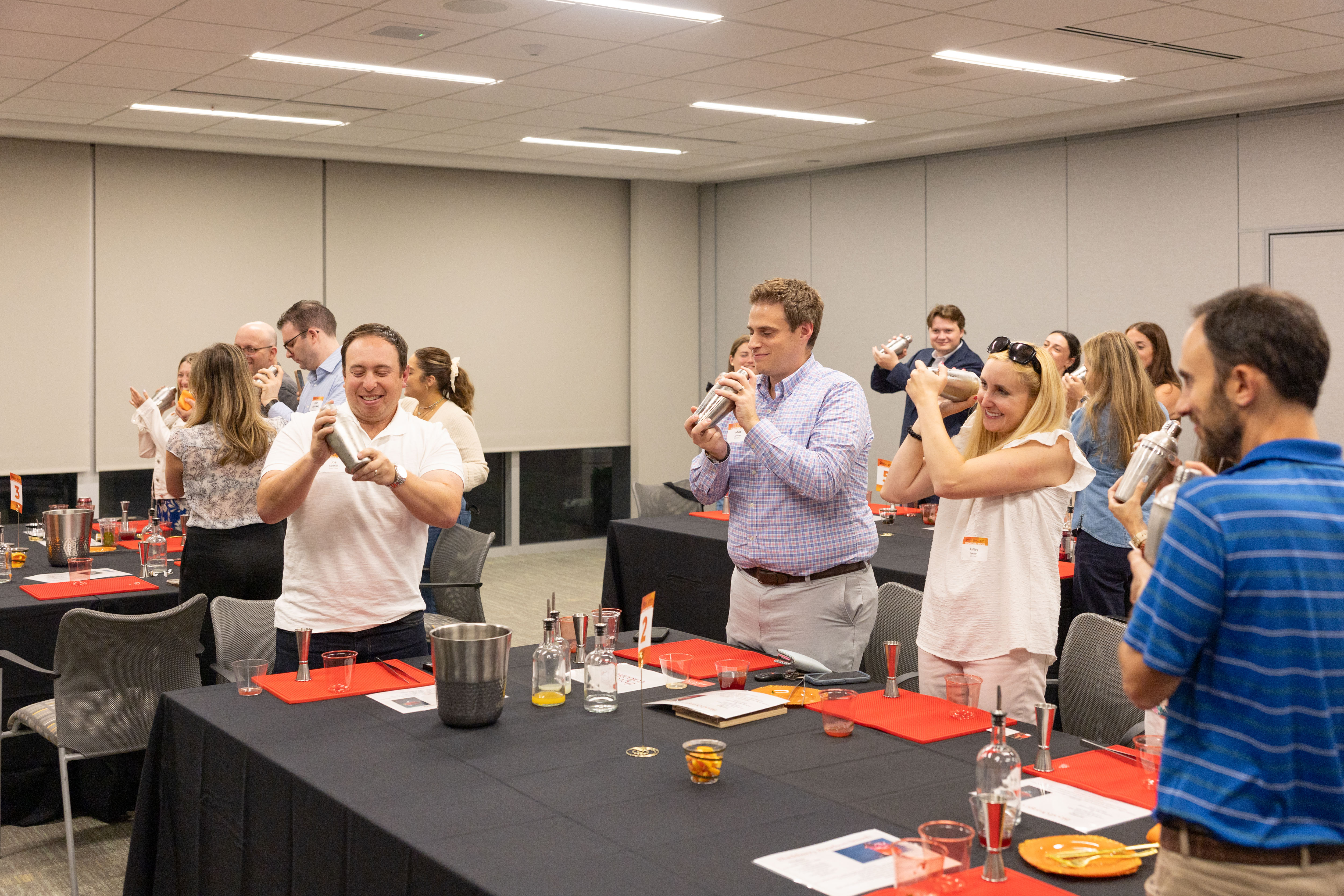 Adults in an office conference room laughing and shaking cocktail shakers during a mixology team-building workshop, tables set with bottles, tools and garnishes.